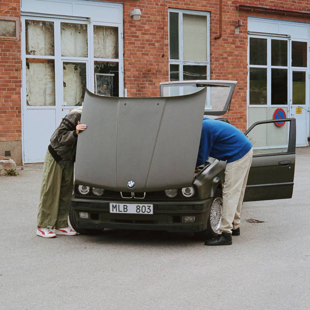 Två personer bakom öppen motorhuv på en svart, trasig bil
